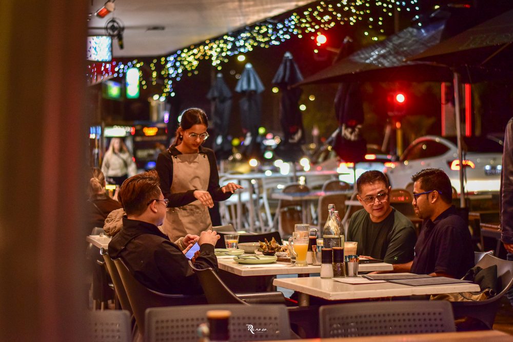 Diners at Mission Bay Cafe on a busy Tāmaki Drive evening, staff serving tables under string lights