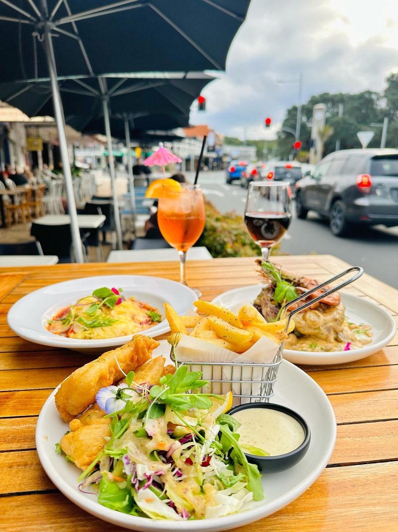 Fish and chips, pasta and cocktails on an outdoor table at Mission Bay Cafe, overlooking Tāmaki Drive